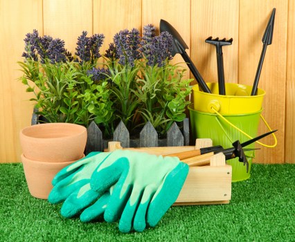 The image displays a neatly arranged gardening setup on a green artificial grass surface, situated against a wooden fence; in the foreground, there is a pair of light terracotta plant pots, followed by a set of green gardening gloves placed on a wooden crate with a black garden tool resting nearby. Behind these are several potted lavender plants with purple flower stalks and green foliage arranged in a row, creating a vibrant contrast against the natural wood background. To the right, a bright yellow plastic bucket holds various gardening tools, including a trowel and a small hand rake, all with black handles. The overall scene suggests a well-organized outdoor space prepared for planting, garden maintenance, or lawn care activities, typical of services provided by Lawn Mowing Barking. The lighting indicates a clear day, emphasizing the fresh and tidy appearance suitable for gardening and landscaping work in the Barking area of East London.