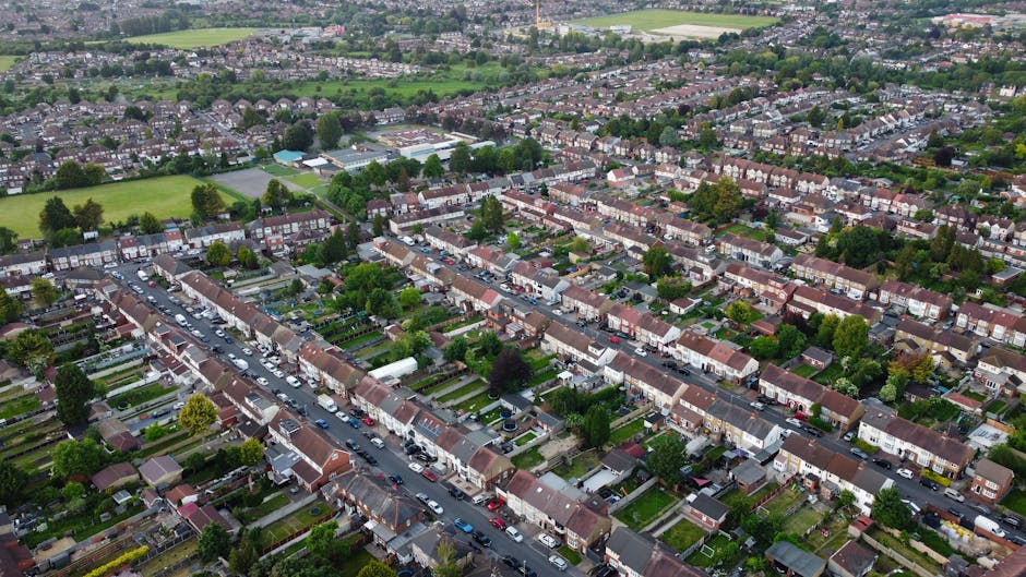 An aerial view of a suburban residential neighborhood showing rows of terraced houses with small front gardens and driveways. The streets are lined with parked cars, and the gardens feature a variety 